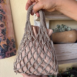Pode incluir: Uma bolsa tipo saco bege com fecho de cordão, envolta numa rede de macramé decorativa de cor taupe. A bolsa apresenta um forro interior liso de cor sólida e duas alças entrançadas que se estendem a partir do topo da estrutura de rede.