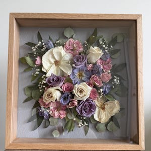 Puede incluir: Un marco de madera cuadrado tipo caja con un arreglo floral prensado. La composición presenta una mezcla de rosas secas en tonos crema, lavanda y rosa, acentuadas con hojas de eucalipto verdes y pequeñas flores blancas de paniculata.