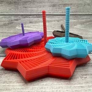 Op de afbeelding: Een verzameling kleurrijke, geometrische 3D-geprinte objecten. De objecten omvatten een rode stervormige basis, een paarse en blauwe top en verticale palen in rood, blauw en bruin. Het ontwerp heeft gelaagde, hoekige patronen.
