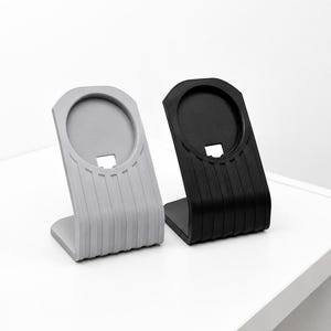 May include: Two phone stands, one light gray and one black, are displayed side-by-side. Each stand has a curved, ribbed design on the front and a circular indentation at the top. The stands are designed to hold a phone or similar device.