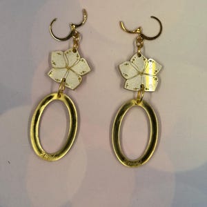Puede incluir: Un par de pendientes colgantes con un aro ovalado de metal dorado suspendido de un dije en forma de flor de cinco pétalos. La flor tiene una superficie plateada reflectante con motas doradas y líneas grabadas. Incluye ganchos dorados.