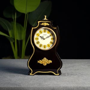 May include: A black mantel clock with gold trim and detailing. The clock face is cream with yellow Roman numerals and black hands. The clock has a curved shape and measures approximately 25 cm in height.