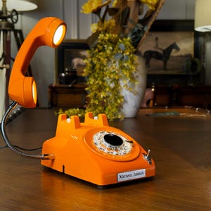 May include: A bright orange desk lamp designed to resemble a vintage rotary telephone. The handset is elevated on a flexible silver neck and functions as a light source. The base features a circular dial plate and a nameplate reading MICHAEL JORDAN.