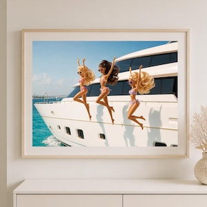 May include: A framed wall art print featuring three dolls in bikinis and sunglasses jumping in the air in front of a large white yacht on blue water. The print is displayed in a light wood frame on a white wall above a white console table.