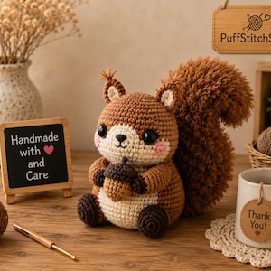May include: A handmade crochet squirrel in shades of brown and cream, holding an acorn. The squirrel has large, expressive eyes and a fluffy tail. A small chalkboard sign reads "Handmade with and Care". A mug with a tag that says "Thank You!" is also present.