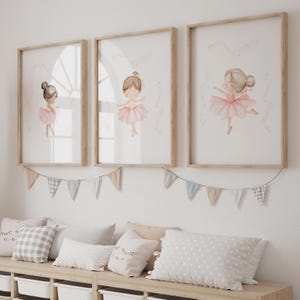 May include: Three framed watercolor prints of ballerinas in pink tutus. The prints are in light wood frames and are hung above a bench with pillows and a banner of triangle flags. The ballerinas have brown hair and are in different dance poses.