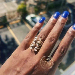 May include: A gold-toned ring with a leaf design, adorned with clear gemstones. The ring is on a hand with blue nail polish. A logo with the letters "TD" is also visible.
