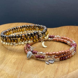 Könnte beinhalten: Drei gewickelte Armbänder mit Perlen. Ein Armband hat Holzperlen in Braun- und Beigetönen. Die anderen beiden Armbänder haben rotbraune, transparente und kupferfarbene Perlen. Jedes Armband hat einen silbernen Anhänger, darunter ein Blatt, ein Mond und ein Baum.