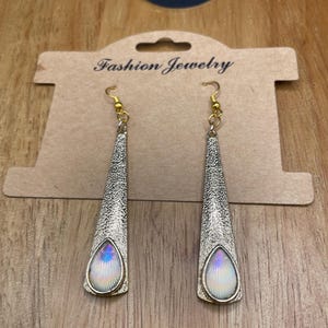 May include: A pair of silver-toned earrings with a textured finish and iridescent teardrop-shaped accents. The earrings hang from gold-colored hooks and are displayed on a brown card that reads "Fashion Jewelry".