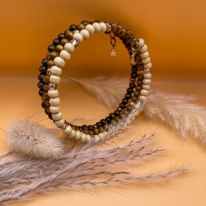 Könnte beinhalten: Ein mehrreihiges Wickelarmband mit abwechselnden Reihen aus glatten cremefarbenen Perlen, dunkelbraunen Tigeraugen-Perlen und kleinen metallischen Zwischenelementen. Das Armband enthält zwei kleine goldfarbene Anhänger, einen sternförmigen und einen mondförmigen.