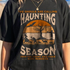 Op de afbeelding: Een zwart t-shirt met een grafische print tegen een oranje zonsondergang. De tekst luidt THE GHOULS ARE CALLING! HAUNTING SEASON JOIN THE HAUNT AND MAKE SOME MEMORIES. De afbeelding bevat een heks, een vleermuis, een boom, een kat en een vogelverschrikker.