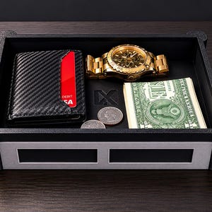 May include: A rectangular black and silver valet tray featuring a carbon fiber textured wallet with a red debit card, a gold-tone analog wristwatch, two silver coins, and a stack of United States dollar bills resting inside the tray compartment.