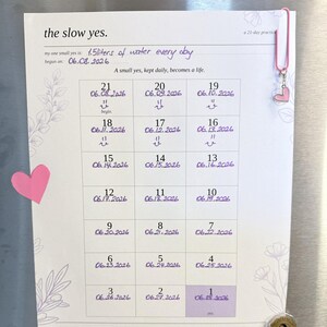 May include: A white paper habit tracker titled the slow yes. a 21-day practice with purple handwritten text stating 1.5 liters of water every day. The grid contains numbered dates from 21 down to 1, decorated with pink heart accents and a pink paperclip.