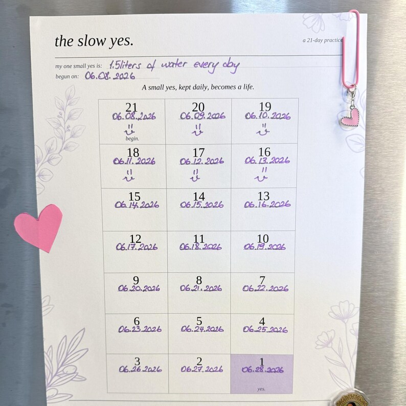 May include: A white paper habit tracker titled the slow yes. a 21-day practice with purple handwritten text stating 1.5 liters of water every day. The grid contains numbered dates from 21 down to 1, decorated with pink heart accents and a pink paperclip.