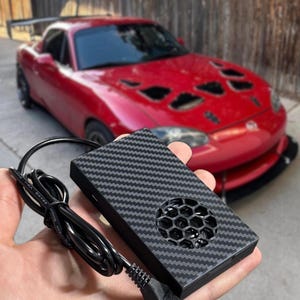 May include: A rectangular black device with a carbon fiber pattern finish and a circular honeycomb-style ventilation grille. The device features an attached black power cable with a standard USB connector extending from the side of the unit.