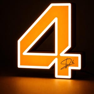 May include: A three-dimensional illuminated number four sign featuring a bright orange face with a white glowing border. The number is constructed with a dark side profile and includes a black handwritten signature on the lower right section.