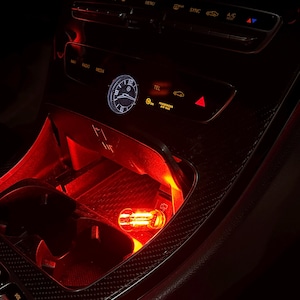 May include: The interior center console of a vehicle featuring a black dashboard with carbon fiber texture, an analog clock, climate control buttons, and a cup holder illuminated by a bright red light, with a small clear light bulb resting inside.