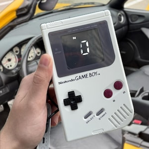 Op de afbeelding: Een witte Nintendo Game Boy handheld spelcomputer met een zwarte richtingspad, twee bordeauxrode A- en B-knoppen en grijze Select- en Start-knoppen. Het scherm toont een groot wit cijfer nul, met de tekst Dot Matrix With Stereo Sound.