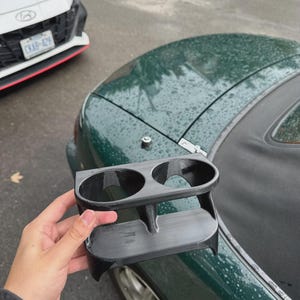 Può includere: Un portabicchieri doppio nero stampato in 3D, dotato di due aperture circolari e di una struttura a strati. L'oggetto presenta una finitura opaca liscia e una base curva progettata per adattarsi alla superficie di una console automobilistica.