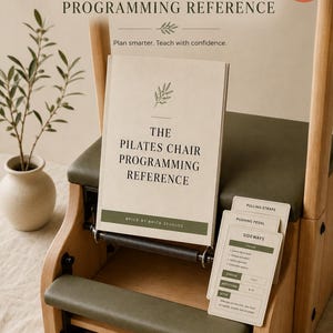 Op de afbeelding: Een Pilates-stoel met een licht houten frame en een olijfgroene zitting. Een handleiding getiteld The Pilates Chair Programming Reference van Brick by Brick Studios rust op de stoel, vergezeld van een stapel instructiekaarten.
