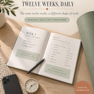 Könnte beinhalten: Ein offener Planer zeigt ein 12-wöchiges tägliches Matte-Übungsprogramm mit Texten wie 20 MINUTES A DAY, TWELVE WEEKS, DAILY, WEEK 1 BUILD THE BASE und DAY 1 BREATH, POSTURE AND CORE. Ein schwarzer Stift, eine runde Uhr und ein Smartphone liegen daneben.