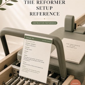 May include: A Pilates reformer machine with a light wood frame and sage green padding. A printed reference card titled The Reformer Setup Reference is attached to the footbar, and a matching manual rests on the carriage. The text includes instructions for footwork, spring settings, and setup.
