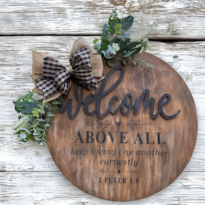 Puede incluir: Un letrero redondo de madera con acabado teñido en marrón oscuro que presenta el texto negro Welcome ABOVE ALL keep loving one another earnestly 1 PETER 4:8. Un lazo de arpillera y cuadros vichy negros se fija con follaje de eucalipto artificial.