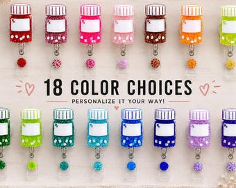 May include: A display of 18 badge reels, each designed as a pill bottle in various colours. The reels feature a white label and a decorative charm. The text reads "18 COLOR CHOICES" and "PERSONALIZE IT YOUR WAY!" on a wooden tray.