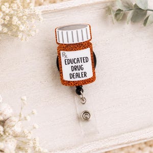 May include: A badge reel featuring a glittery orange prescription bottle shape with a white cap. The label displays the Rx symbol and the black text "EDUCATED DRUG DEALER" in a sans-serif font. The reel includes a clear plastic strap and a metal snap.
