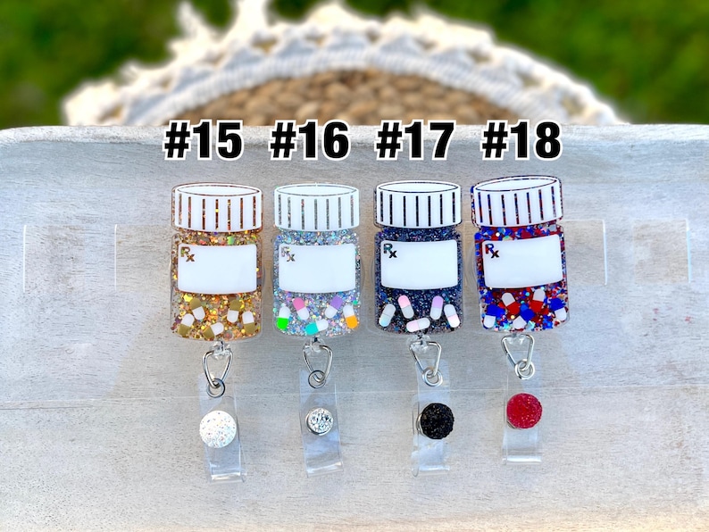 May include: Four prescription bottle-shaped badge reels with glitter and decorative accents. Each reel has a different color scheme, including gold, silver, navy, and red. The reels are labeled with the numbers #15, #16, #17, and #18.