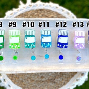 May include: A collection of colorful, glittery prescription bottle-shaped badge reels. Each reel is a different color, including green, blue, and purple, with a white label and the letters "Rx". The reels are numbered from #8 to #14.