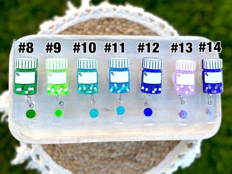 May include: A collection of colorful, glittery prescription bottle-shaped badge reels. Each reel is a different color, including green, blue, and purple, with a white label and the letters "Rx". The reels are numbered from #8 to #14.