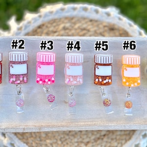May include: A collection of seven prescription bottle-shaped badge reels in various colors. Each reel has a white label, a glittery base, and a matching glitter accent. The bottles are numbered from one to seven.