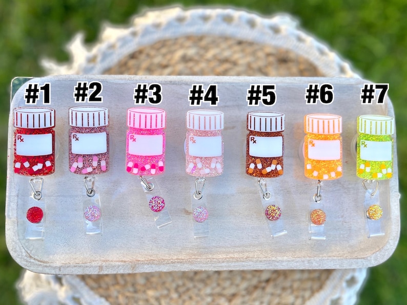 May include: A collection of seven prescription bottle-shaped badge reels in various colors. Each reel has a white label, a glittery base, and a matching glitter accent. The bottles are numbered from one to seven.