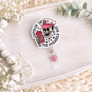 May include: A white badge reel with a skull wearing a pink captain's hat and holding a red cup. The text "CAPTAIN OF THE STRUGGLE BUS" surrounds the skull. The reel has a pink glitter accent.