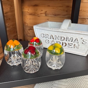 May include: Three clear glass paperweights with faceted bases, each containing embedded miniature fruit shapes including orange, red, and yellow pieces nestled in green foliage. A white wooden basket with the text GRANDMA'S GARDEN is visible.