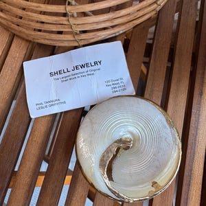 May include: A circular shell ornament with a pearlescent white centre and a gold-toned metallic rim. Beside it lies a white business card printed with the text: SHELL JEWELRY, The Largest Selection of Original Shell Work in Key West, 120 Duval St.