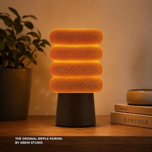 May include: A cylindrical table lamp with a dark gray base and a glowing orange, ribbed, fabric-covered shade. The lamp features four distinct horizontal rounded sections. Text at the bottom reads: THE ORIGINAL RIPPLE PAIRING BY ARDIN STUDIO.