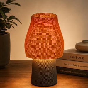 May include: A small, modern table lamp with an orange fabric shade and a dark gray base. The lamp is illuminated, casting a warm glow. The lamp is sitting on a wooden surface.