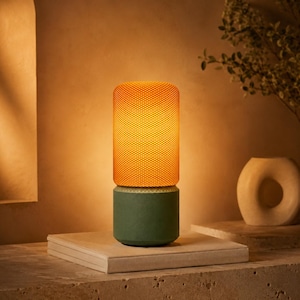Puede incluir: Una lámpara de mesa moderna con una pantalla cilíndrica naranja y una base verde. La pantalla tiene un patrón texturizado en forma de rejilla, y la base es verde mate. La lámpara está encendida, emitiendo un brillo cálido y acogedor.