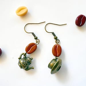 May include: A pair of dangle earrings featuring bronze-toned metal charms shaped like a teapot and a teacup, each accented with a single glossy brown coffee bean bead suspended from a bronze-colored hook wire, surrounded by loose coffee beans.