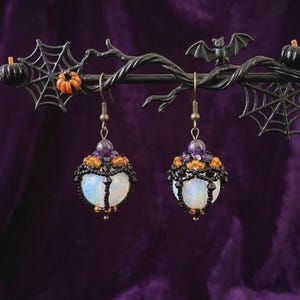 May include: A pair of drop earrings featuring spherical white iridescent beads encased in black beaded netting, accented with small orange flower-shaped beads and purple round beads at the top, suspended from bronze-toned metal earring hooks.