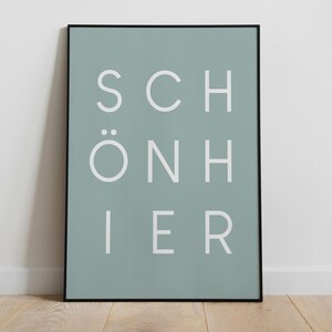 Puede incluir: Un póster rectangular enmarcado en un borde negro fino con un fondo verde salvia y el texto blanco SCHÖN HIER dispuesto en tres filas. La tipografía es limpia, sin remates y centrada, creando una composición visual equilibrada.