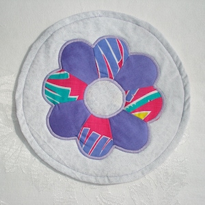 May include: A round, quilted coaster featuring a large flower design. The flower has purple petals and a center with pink, teal, and yellow accents. The coaster's outer ring is light gray with a subtle swirl pattern.