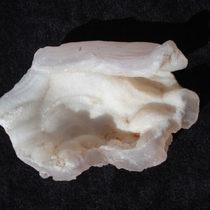 May include: A close-up of a white, irregularly shaped mineral specimen. The surface has a layered, textured appearance with subtle variations in tone. The mineral sits against a dark background, highlighting its form and detail.
