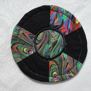 May include: Circular coaster with a black and colorful abstract design. The coaster is made of fabric and features a central circle with a swirling pattern of green, orange, and purple. The outer sections are black.