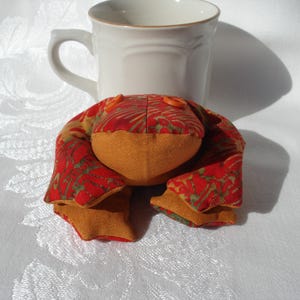May include: A handmade fabric frog with a red and green patterned body and orange accents. The frog is positioned in front of a white ceramic mug with a curved handle. The frog's eyes are small orange buttons.