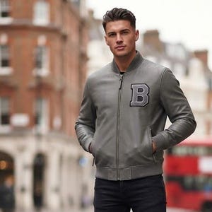 May include: A gray bomber jacket with a ribbed collar, cuffs, and hem, featuring a large letter B patch on the left chest. The jacket has a full-length front zipper and is paired with slim-fit black denim jeans and plain white leather sneakers.