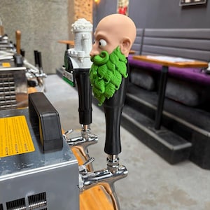May include: A beer tap handle featuring a sculpted head with a bald pate and a prominent green beard made of textured leaves. The handle is mounted on a chrome faucet attached to a silver metal cooling unit, positioned next to a white castle-shaped tap.