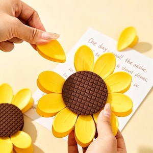 Puede incluir: Clips amarillos con forma de girasol y centros marrones. Los clips están siendo sostenidos por la mano de una persona. Un trozo de papel con texto manuscrito está en el fondo. Los clips son de color amarillo brillante.